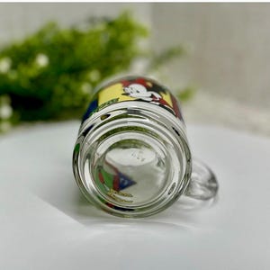 Peut inclure: Mug en verre transparent avec une poign&eacute;e, pr&eacute;sentant un dessin de dessin anim&eacute; color&eacute;. Le motif comprend Mickey Mouse dans un sch&eacute;ma de couleurs jaune, rouge et noir. Le mug est sur une surface blanche.