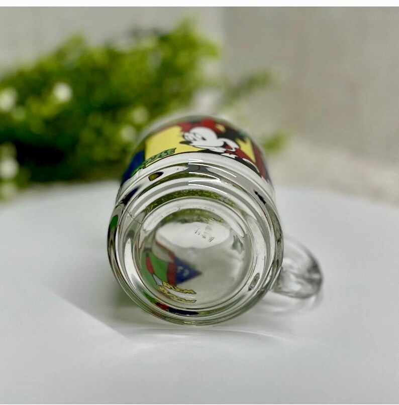 Peut inclure: Mug en verre transparent avec une poign&eacute;e, pr&eacute;sentant un dessin de dessin anim&eacute; color&eacute;. Le motif comprend Mickey Mouse dans un sch&eacute;ma de couleurs jaune, rouge et noir. Le mug est sur une surface blanche.