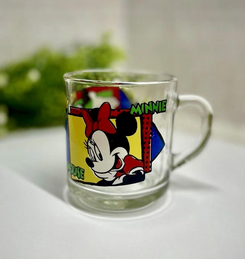 Peut inclure: Mug en verre transparent avec Minnie Mouse. Le mug a une anse et un graphisme color&eacute; de Minnie Mouse avec un n&oelig;ud rouge, des gants rouges et un fond jaune. Le mot "MINNIE" est imprim&eacute; sur le mug.