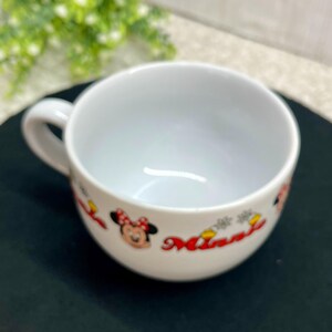 K&ouml;nnte beinhalten: Wei&szlig;e Keramik-Suppentasse mit Henkel. Die Tasse zeigt ein Minnie Mouse-Design mit roter Schleife und dem Namen "Minnie" in roter Schrift. Die Tasse steht auf einer schwarzen Oberfl&auml;che.