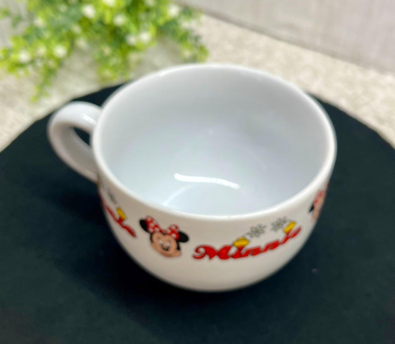 K&ouml;nnte beinhalten: Wei&szlig;e Keramik-Suppentasse mit Henkel. Die Tasse zeigt ein Minnie Mouse-Design mit roter Schleife und dem Namen "Minnie" in roter Schrift. Die Tasse steht auf einer schwarzen Oberfl&auml;che.