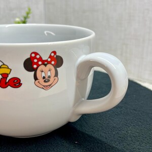 K&ouml;nnte beinhalten: Wei&szlig;e Keramiktasse mit Minnie Mouse und einer roten Schleife mit Tupfen. Die Tasse hat einen gro&szlig;en, gebogenen Henkel und eine weite &Ouml;ffnung. Das Wort "Minnie" ist rot und gelb aufgedruckt.
