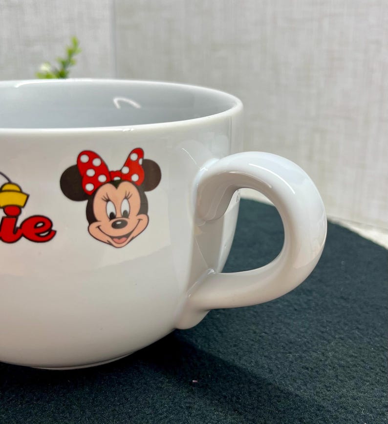 K&ouml;nnte beinhalten: Wei&szlig;e Keramiktasse mit Minnie Mouse und einer roten Schleife mit Tupfen. Die Tasse hat einen gro&szlig;en, gebogenen Henkel und eine weite &Ouml;ffnung. Das Wort "Minnie" ist rot und gelb aufgedruckt.