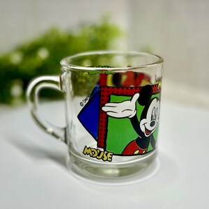 Peut inclure: Mug en verre transparent avec une poign&eacute;e, repr&eacute;sentant Mickey Mouse. Le mug pr&eacute;sente un motif color&eacute; de Mickey Mouse avec le mot "MOUSE" en bas. Le design comprend des couleurs rouge, vert, bleu et jaune.