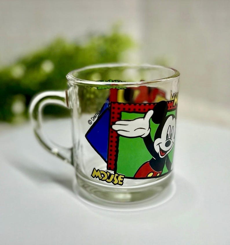 Peut inclure: Mug en verre transparent avec une poign&eacute;e, repr&eacute;sentant Mickey Mouse. Le mug pr&eacute;sente un motif color&eacute; de Mickey Mouse avec le mot "MOUSE" en bas. Le design comprend des couleurs rouge, vert, bleu et jaune.