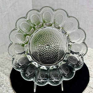 May include: Clear glass serving dish with a scalloped edge and a flower-like design. The center of the dish has a textured pattern, and the outer edge features oval-shaped indentations. The dish is supported by a clear stand.