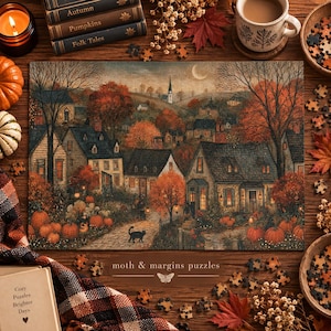 Pode incluir: Um quebra-cabeça retangular com uma cena de vila outonal, casas, abóboras laranjas, um gato preto e uma lua crescente. O texto 'moth & margins puzzles' está impresso abaixo da imagem, acompanhado por uma pequena ilustração de uma mariposa.