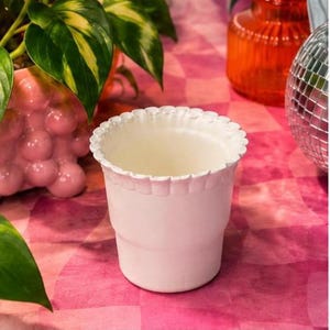May include: A white, scalloped-edge planter pot. The pot is empty and sits on a pink and red patterned surface. Other decorative items, including a disco ball and a pink bubble-shaped vase, are in the background.