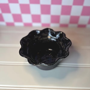 May include: A small, glossy black ceramic bowl with a distinctively wavy, scalloped rim. The bowl features a smooth, reflective surface and a deep, rounded basin, showcasing a hand-formed, organic shape with irregular, undulating edges.