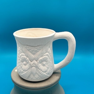 May include: White ceramic mug with an owl face design. The mug has a large handle and is sitting on a gray pedestal against a blue background. The owl's features are detailed and embossed on the mug's surface.