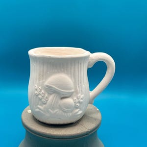 Op de afbeelding: Een witte keramische mok met een paddestoelontwerp. De mok heeft een oor en staat op een grijze sokkel tegen een blauwe achtergrond. De buitenkant van de mok heeft een gestructureerd, verticaal patroon.