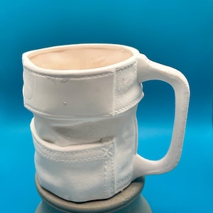 May include: A white ceramic mug with a denim-style design. The mug has a handle and is supported by a grey pedestal. The mug's design includes stitched details, resembling pockets and seams of denim jeans.