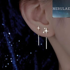 May include: Silver star-shaped stud earrings and dangling bar earrings. The earrings are worn on an ear, with a dark blue background. The word "Nebulae" is visible in the upper right corner.