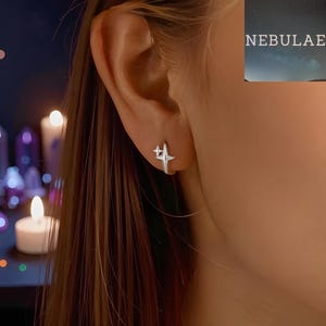 Op de afbeelding: Een zilveren oorbel in de vorm van een ster wordt op een oor gedragen. De oorbel heeft een vierpuntig sterontwerp. Het woord "NEBULAE" is zichtbaar in de rechterbovenhoek. De achtergrond is wazig, met zachte verlichting van kaarsen.