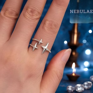 Puede incluir: Un anillo plateado con tres diseños en forma de estrella. El anillo se lleva en un dedo, con las estrellas dispuestas para crear una estética celestial. La palabra "NEBULAE" es visible en el fondo.