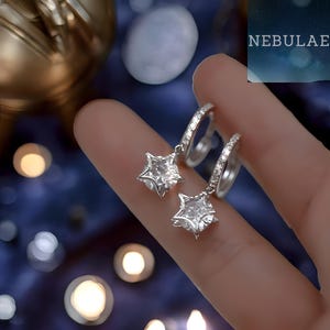 May include: A pair of silver hoop earrings with star-shaped pendants. The earrings feature clear, faceted gemstones and are displayed against a blurred, dark blue background with the word "NEBULAE".