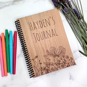 May include: A wooden journal with a spiral binding. The cover is engraved with the text "HAYDEN'S JOURNAL" and a floral design. The journal is on a white surface with a bouquet of lavender and a green plant.