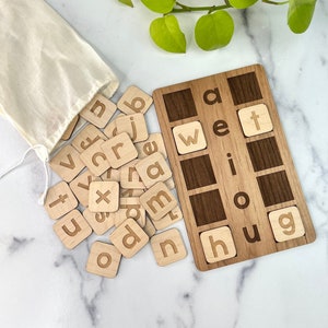 May include: Wooden letter tiles in a white drawstring bag, with a wooden board with the words "a wet i o hug" on it. The tiles are scattered around the board.