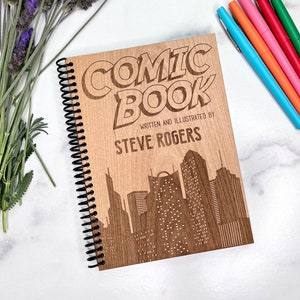 May include: A wooden spiral-bound notebook with the words "COMIC BOOK" and "WRITTEN AND ILLUSTRATED BY STEVE ROGERS" engraved on the cover. The cover also features a cityscape silhouette. The notebook is surrounded by colorful pens and lavender.