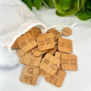 May include: A wooden bingo game set with a drawstring bag. The set includes numerous square wooden tiles with engraved letters and numbers. A small wooden tag with the word "BINGO" is attached to the bag.
