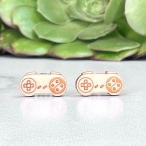 May include: A pair of small, white, plastic earrings shaped like a classic video game controller. The controller is brown with a cross, circle, triangle, and square buttons.