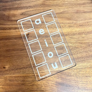 May include: Clear acrylic learning tool with five squares and the letters a, e, i, o, and u printed on it.