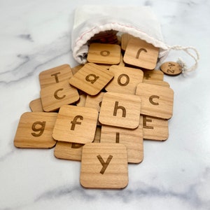May include: A set of wooden letter tiles in a white drawstring bag. The tiles are scattered on a white marble surface. The tiles are made of light brown wood and have the letters of the alphabet engraved on them. The bag has a small tag that says "A-Z".