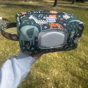 May include: A dark green wipe dispenser with a handle, decorated with a jungle animal print. The design includes elephants, zebras, giraffes, and monkeys. A white dispenser lid is visible. The label reads "Maddy Made".