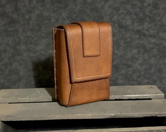 May include: A rectangular brown leather pouch featuring a vertical flap closure with a centered strap detail. The pouch has visible stitched edges along the side and a smooth, matte finish, constructed with a structured design for secure storage.