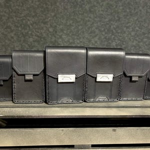 Peut inclure: Six pochettes en cuir noir de différentes tailles sont disposées sur une surface en bois. Chaque pochette est dotée d'une fermeture à rabat avec un fermoir en métal argenté. Les pochettes sont cousues avec un fil contrastant.