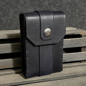 May include: A black leather case with a flap closure and a silver snap. The case is rectangular with a vertical strap and a stitched edge. The background is a dark, textured surface.