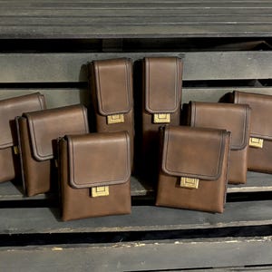 May include: A collection of brown leather cases with a rectangular shape and a flap closure. Each case has a gold-colored clasp. The cases are arranged on a dark wooden surface.