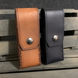 May include: Two leather knife sheaths, one brown and one black, with a snap closure and stitching detail.