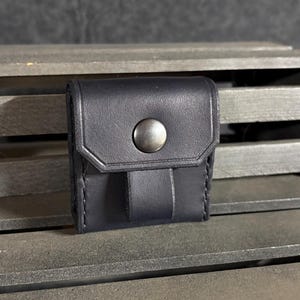 May include: A small, square black leather pouch featuring a single flap closure secured by a centered, circular metallic snap button. The pouch has visible stitching along the edges and a vertical leather loop attached to the front flap.