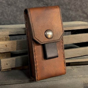 May include: A brown leather case with a flap closure and a metal button. The case has a stitched edge and a black leather strap.