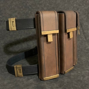May include: A brown leather leg holster with two pouches and gold metal accents. The holster has a black adjustable strap with a buckle.