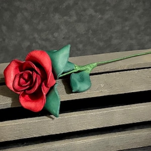May include: A handcrafted red rose with green leaves and stem, lying on a wooden surface. The rose's petals are a vibrant red, and the leaves and stem are a deep green. The rose is a symbol of love and beauty.