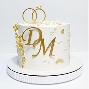 Könnte beinhalten: Eine weiße Torte, dekoriert für eine Hochzeit oder Verlobung. Die Torte hat goldene Initialen "D" und "M", goldene Blattakzente und einen goldenen Ring-Tortentopper mit einem Diamanten.