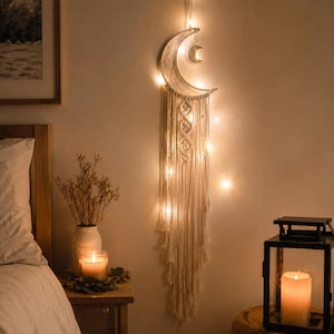 May include: A crescent moon wall hanging made of off-white macrame cord, featuring a smaller metal moon charm suspended within the frame. The piece is illuminated by integrated warm white string lights that extend down the long, knotted fringe.