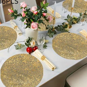 May include: A set of 36 round gold-toned placemats with a textured, marbled pattern. The placemats are arranged on a white table alongside a white vase filled with pink roses, clear glass stemware, white cloth napkins, and gold candle holders.
