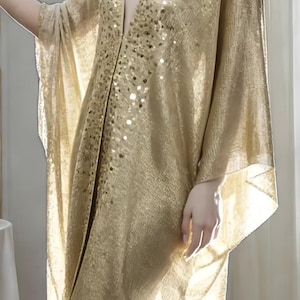 May include: A gold-colored, semi-sheer kaftan featuring a deep V-neckline, wide kimono-style sleeves, and a front panel embellished with shimmering gold sequins. The garment has a textured, metallic finish and a fringed hemline at the bottom edge.