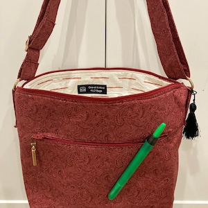 May include: A red paisley-patterned shoulder bag with a gold zipper and a black tassel. The bag has a front pocket and a green pen is in the pocket. The interior is white with red stripes. The label reads "One-of-A-Kind KLO Bags."