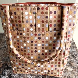 Op de afbeelding: Een bruine tote bag met een herhaald patroon van kleurrijke stippen. De tas heeft een voorvak en gewatteerde handvatten. De binnenbekleding is effen roestkleurig.