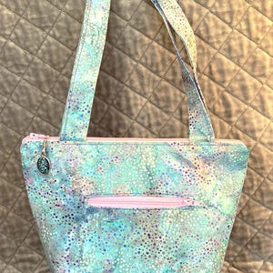 Op de afbeelding: Een tote bag met een lichtblauw, groen en paars gespikkeld patroon. De tas heeft een roze rits en een bijpassend roze ritsvak aan de voorkant. De handvatten hebben hetzelfde gespikkelde patroon als de tas. Een kleine bedel hangt aan de rits.