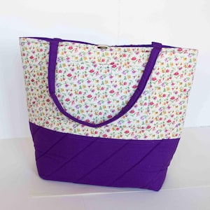 May include: A quilted tote bag with a floral pattern on a white background and a solid purple bottom. The bag has purple handles and a metal clasp. The floral pattern includes pink, yellow, and purple flowers.