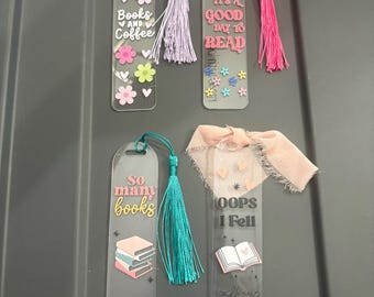 Könnte beinhalten: Fünf transparente Acryl-Lesezeichen mit Zitaten über das Lesen und dekorativen Quasten. Die Lesezeichen zeigen Sätze wie "Books and Coffee" und "It's a good day to read". Jedes Lesezeichen hat ein anderes Design, darunter Blumen und Herzen.