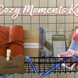 May include: A Cozy Moments Kit displayed in a black wire basket. The kit includes an orange leather journal with leaf detailing, a small card, stickers, a pair of socks, and a tag that reads "Cozy moments". A blue and white ribbon decorates the basket.