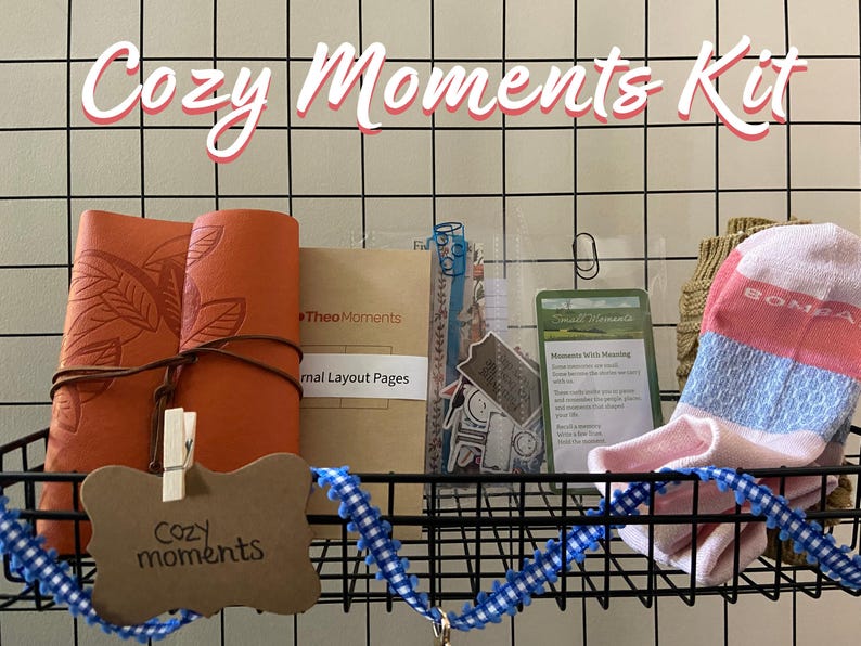 May include: A Cozy Moments Kit displayed in a black wire basket. The kit includes an orange leather journal with leaf detailing, a small card, stickers, a pair of socks, and a tag that reads "Cozy moments". A blue and white ribbon decorates the basket.