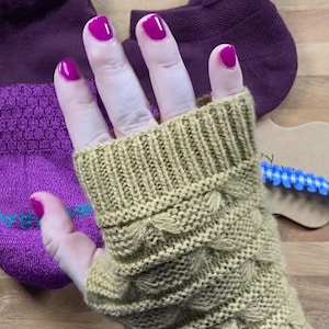 May include: A hand wearing a knitted, olive green fingerless glove with a textured, scalloped design. The hand has bright pink nail polish. A pair of purple socks are in the background.