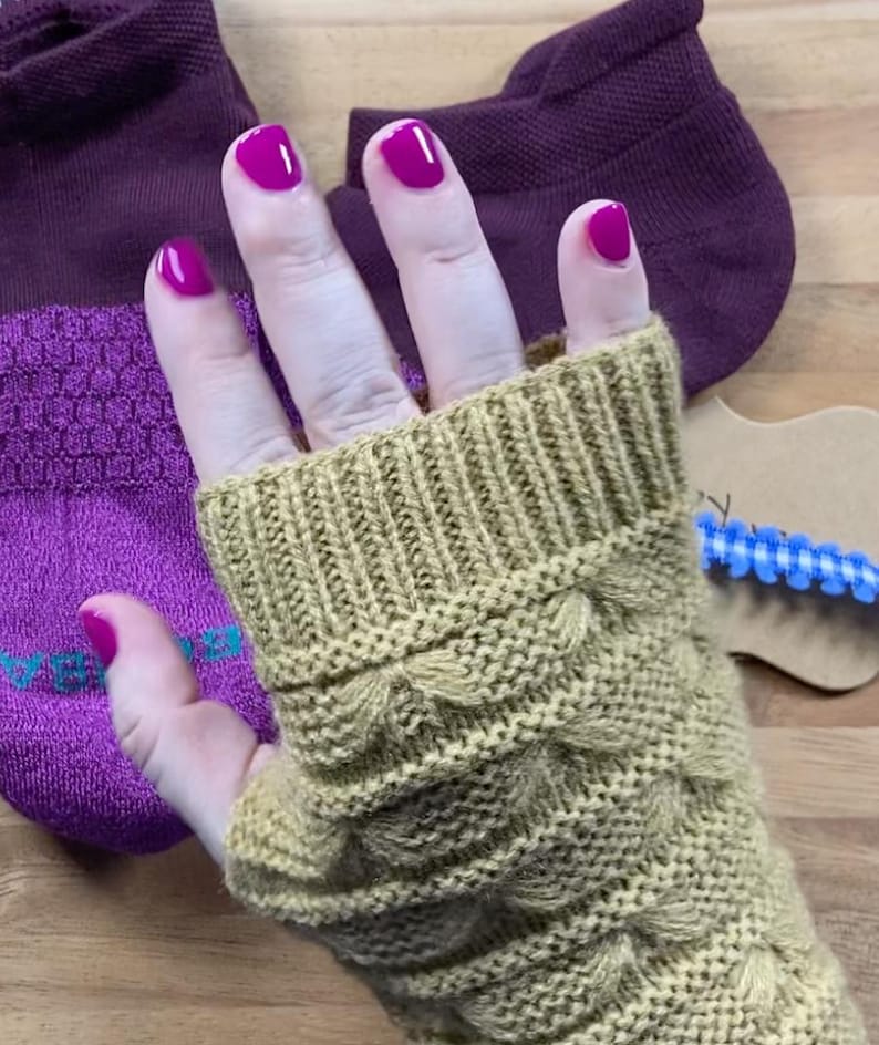 May include: A hand wearing a knitted, olive green fingerless glove with a textured, scalloped design. The hand has bright pink nail polish. A pair of purple socks are in the background.